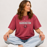 A woman sitting cross-legged wearing a maroon short-sleeve graphic T-shirt that reads 'dance team BARRINGTON' with a horseshoe logo, paired with light blue jeans and white sneakers, against a plain light gray background.