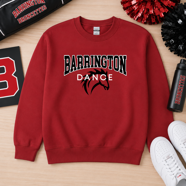 A red crewneck sweatshirt with 'BARRINGTON DANCE' and a horse graphic printed on the front, laid flat on a light wood surface with cheerleader pom-poms, a water bottle, and white sneakers around it.