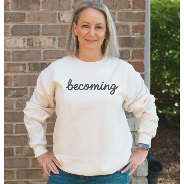 A woman with blonde hair stands in front of a brick wall wearing a cream-colored crewneck sweatshirt with the word 'becoming' printed in black script across the chest, paired with blue jeans.