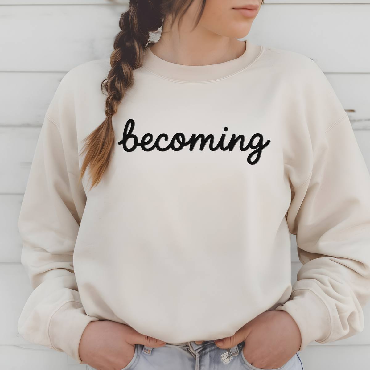 A woman wearing a cream-colored crewneck sweatshirt with the word 'becoming' printed in black script across the chest, paired with light blue jeans, set against a white wood-paneled background.