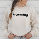 A woman wearing a cream-colored crewneck sweatshirt with the word 'becoming' printed in black script across the chest, paired with light blue jeans, set against a white wood-paneled background.