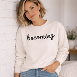 A woman with blonde hair leans against a white brick wall wearing a cream-colored crewneck sweatshirt with the word 'becoming' printed in black script across the chest, paired with blue jeans.