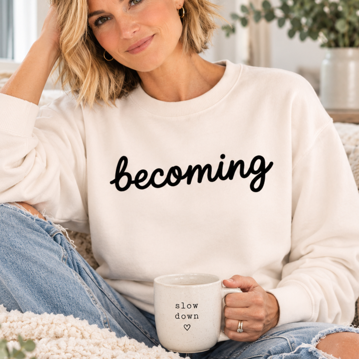 A woman sits cross-legged on a cozy cream-colored couch, wearing a white crewneck sweatshirt with the word 'becoming' printed in black script across the chest. She is holding a white ceramic mug that reads 'slow down' with a small heart.