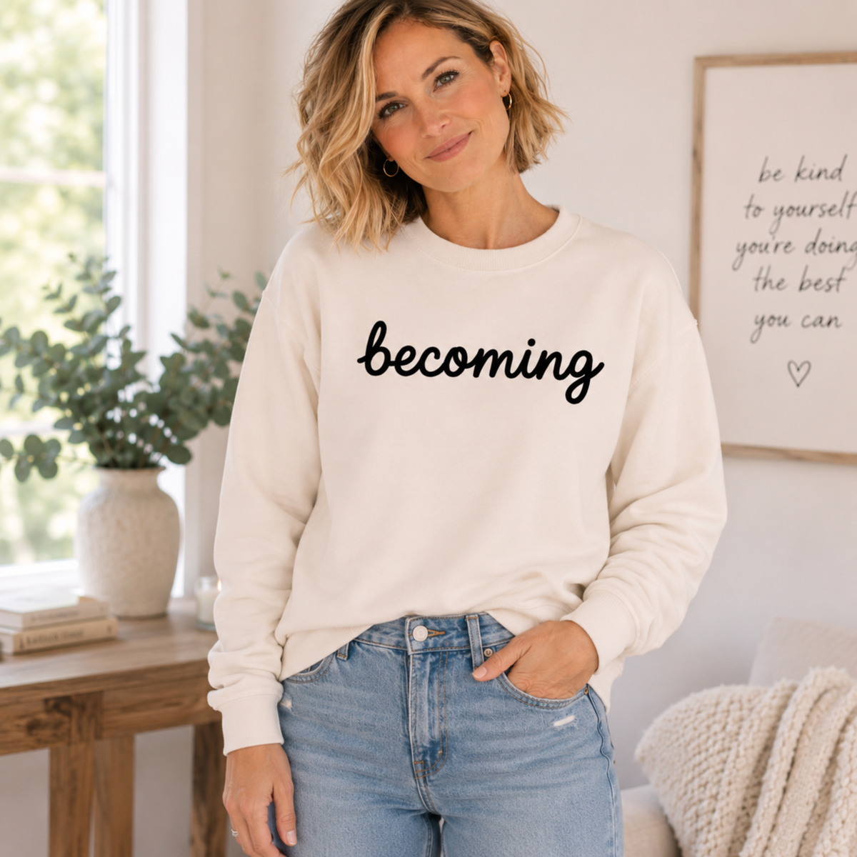 A woman wearing a cream-colored crewneck sweatshirt with the word 'becoming' printed in black script across the chest, paired with light blue jeans in a bright, modern room.