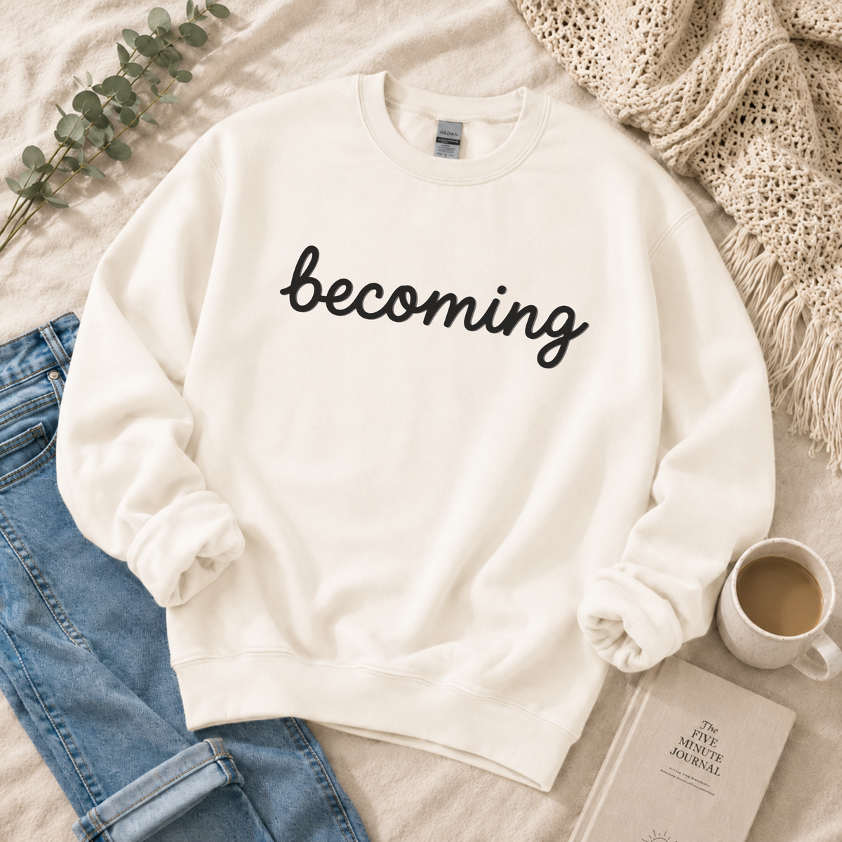 An off-white crewneck sweatshirt with the word 'becoming' printed in black cursive across the chest, styled in a cozy flat-lay arrangement with blue jeans, a cup of coffee, a book, and a knit blanket.