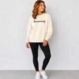 A woman modeling a cream-colored crewneck sweatshirt with the word 'becoming' embroidered in black script across the chest, paired with black leggings and white sneakers against a light gray studio background.