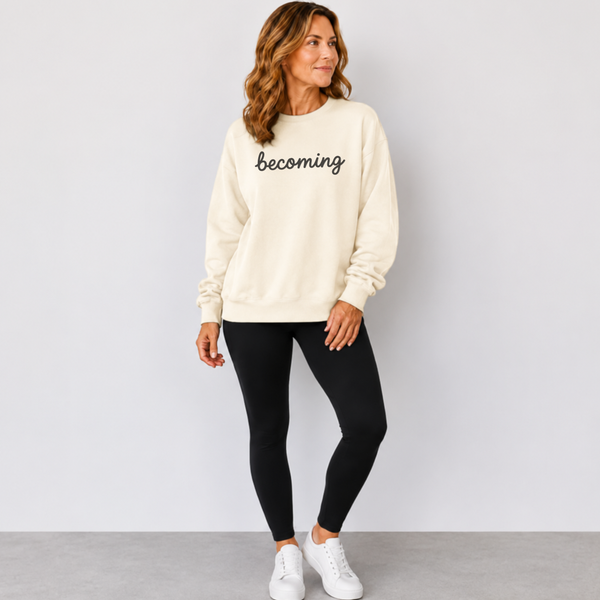 A woman modeling a cream-colored crewneck sweatshirt with the word 'becoming' embroidered in black script across the chest, paired with black leggings and white sneakers against a light gray studio background.