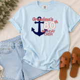 A light blue graphic T-shirt laid flat on a beige fabric surface, styled with denim shorts, brown sandals, and eucalyptus leaves. The shirt has a large anchor graphic and the text 'Grandma's 80th Birthday Crew' in pink and navy.