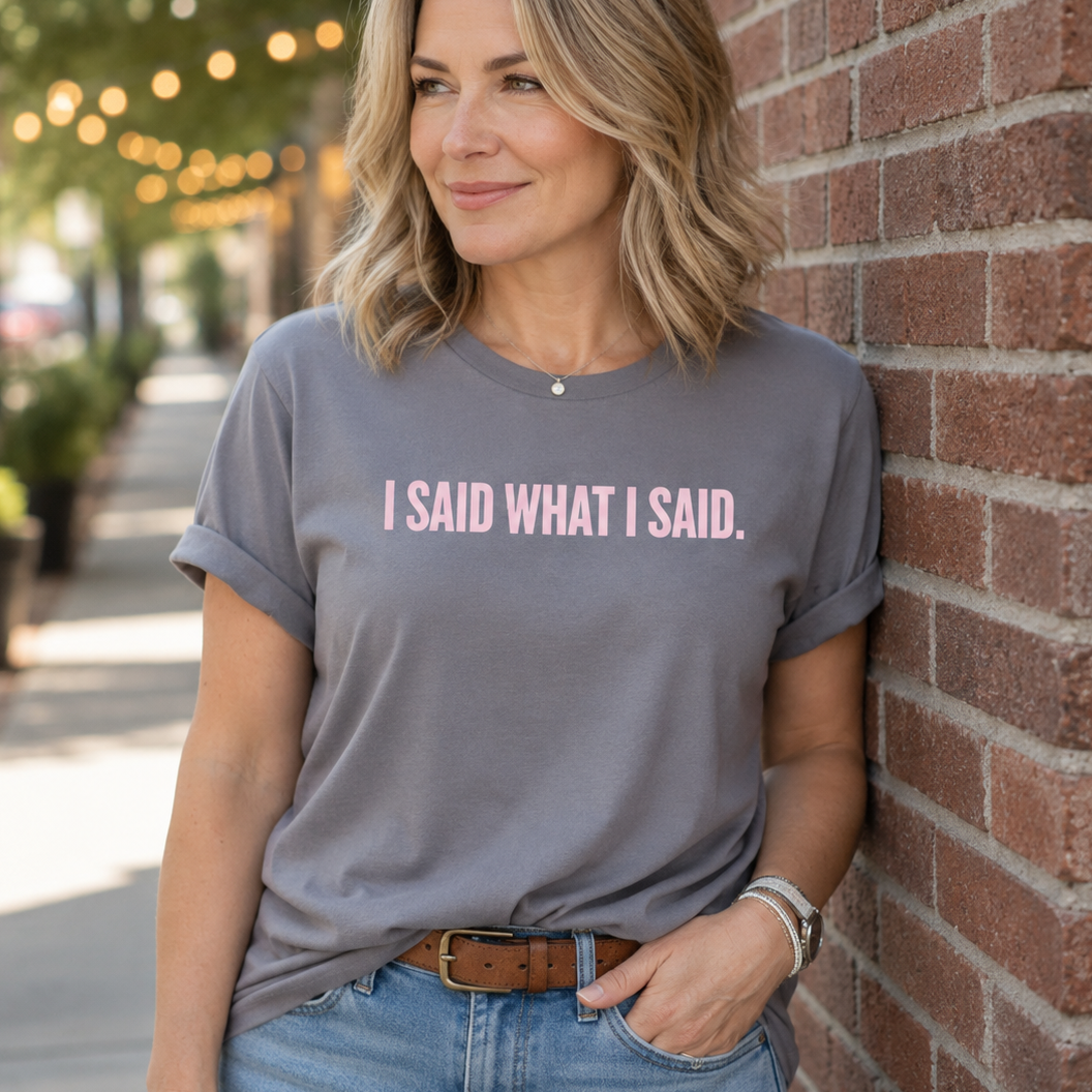 A woman wearing a heather gray t-shirt with the text 'I SAID WHAT I SAID.' in pink, leaning against a red brick wall outdoors.