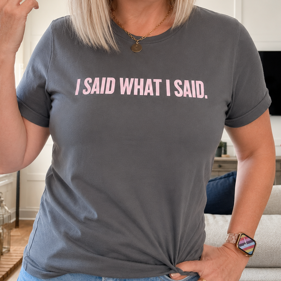 A woman wearing a dark gray t-shirt with the text 'I SAID WHAT I SAID.' printed in light pink across the chest, standing in a living room setting.