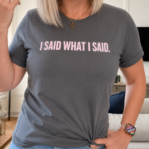 A woman wearing a dark gray t-shirt with the text 'I SAID WHAT I SAID.' printed in light pink across the chest, standing in a living room setting.
