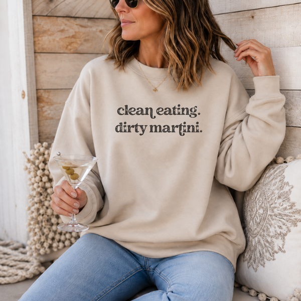 A woman wearing a beige crewneck sweatshirt with the text 'clean eating. dirty martini.