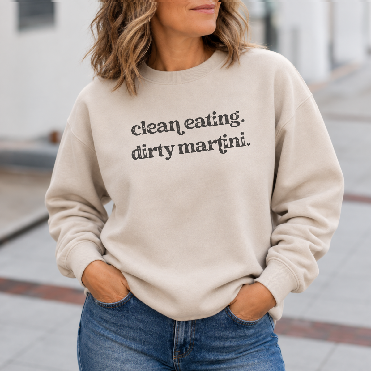 A woman wearing a beige crewneck sweatshirt with the text 'clean eating. dirty martini.