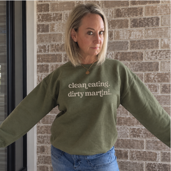 A woman wearing an olive green crewneck sweatshirt with the text 'clean eating. dirty martini.