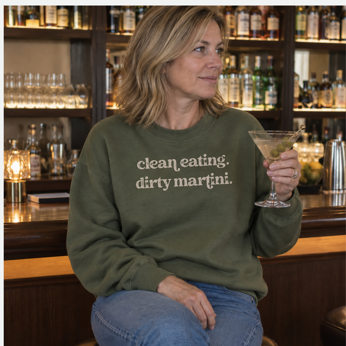 A woman sitting at a bar wearing an olive green crewneck sweatshirt with the text 'clean. eating.