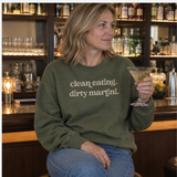 A woman sitting at a bar wearing an olive green crewneck sweatshirt with the text 'clean. eating.