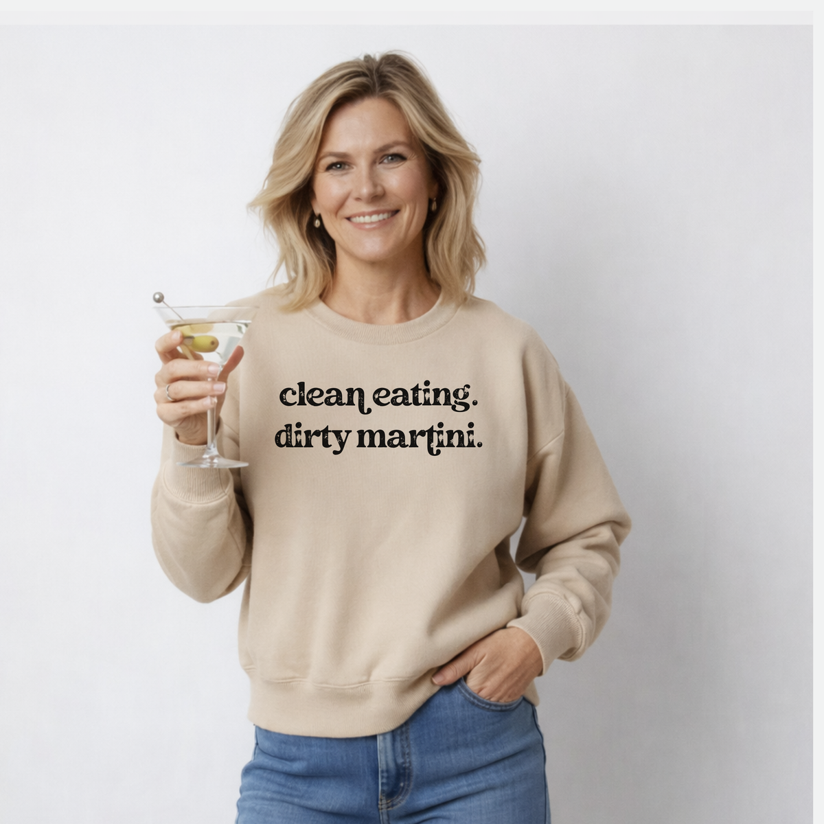 Woman wearing a beige sweatshirt with 'clean eating. dirty martini.' text, holding a martini glass.
