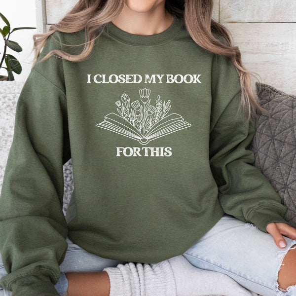Person wearing a green sweatshirt with a book and plant design and text that reads "I CLOSED MY BOOK FOR THIS."