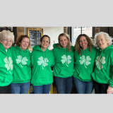 A group of 6 woman wearing green sweatshirts with a large lighter green shamrock in the middle.