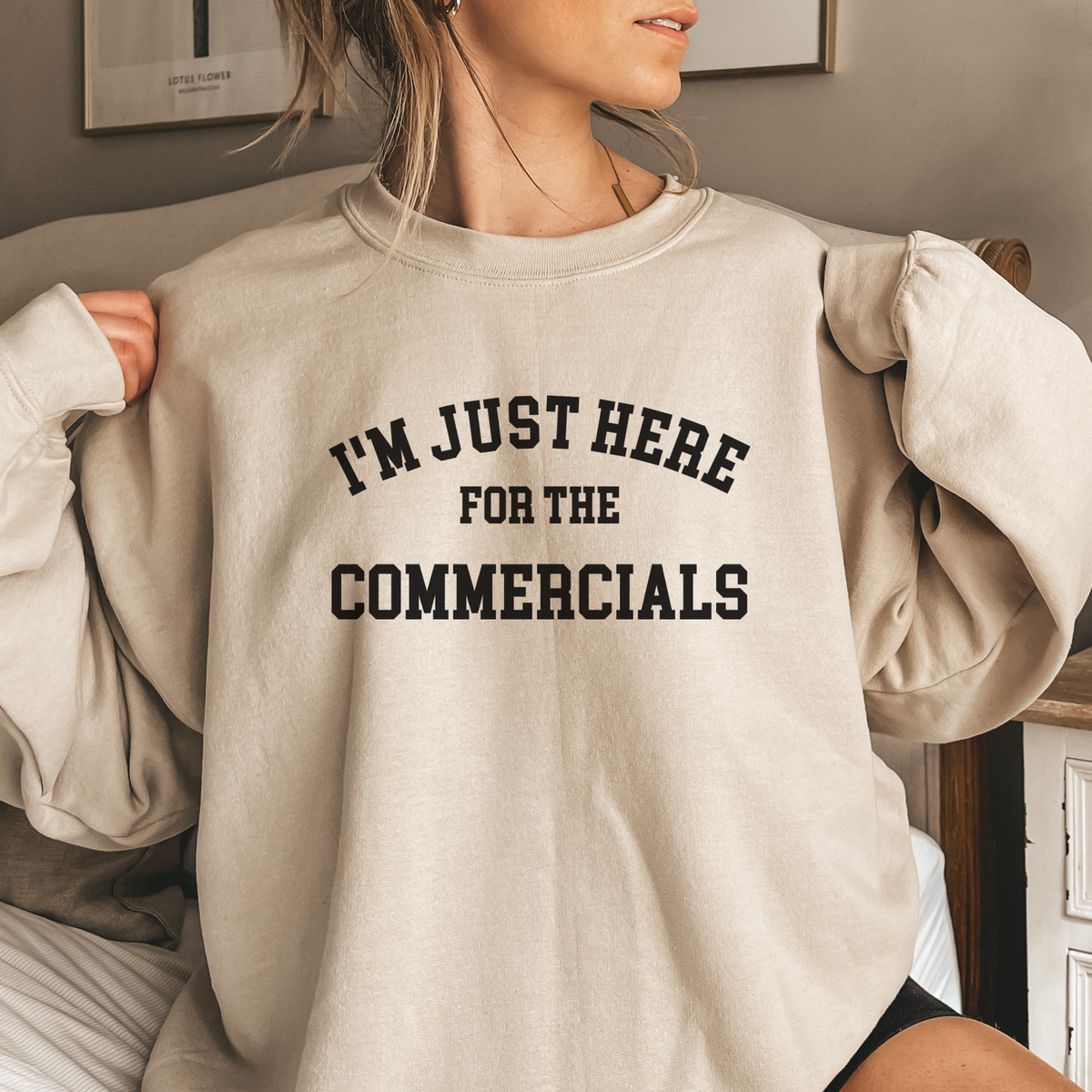 Person wearing an oversized beige sweatshirt with text 'I'm just here for the commercials' in a casual setting.