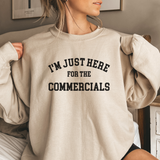 Person wearing an oversized beige sweatshirt with text 'I'm just here for the commercials' in a casual setting.