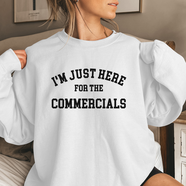 Person wearing a white sweatshirt with text 'I'm just here for the commercials' in a casual setting.