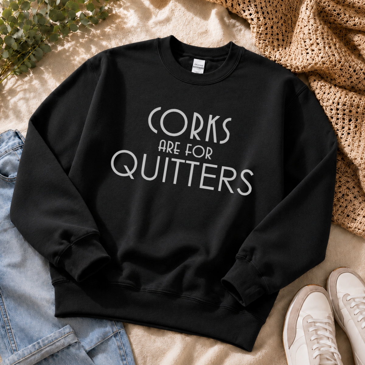 A black crewneck sweatshirt with the text 'CORKS ARE FOR QUITTERS' printed in white across the chest, styled in a flat lay with jeans, a woven blanket, and white sneakers.