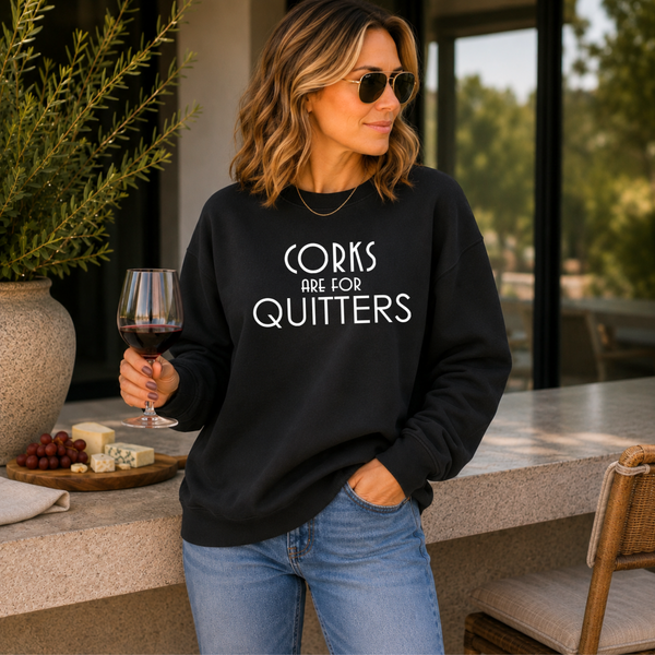 A woman wearing a black crewneck sweatshirt with the white printed text 'CORKS ARE FOR QUITTERS' stands in a modern patio setting holding a glass of red wine.