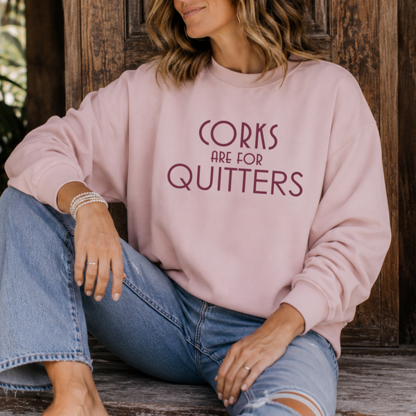 A woman wearing a dusty pink crewneck sweatshirt with the text 'CORKS ARE FOR QUITTERS' printed in dark maroon on the front, paired with blue jeans, sitting against a rustic wooden background.