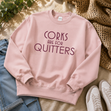 A light pink crewneck sweatshirt with the text 'CORKS ARE FOR QUITTERS' printed in dark purple across the chest, styled in a flat lay with blue jeans, white sneakers, a woven blanket, and green leaves on a beige fabric background.