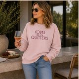 A woman wearing a light pink crewneck sweatshirt with the text 'CORKS ARE FOR QUITTERS' in dark pink lettering, paired with blue jeans and sunglasses, holding a glass of rosé wine in an outdoor patio setting.