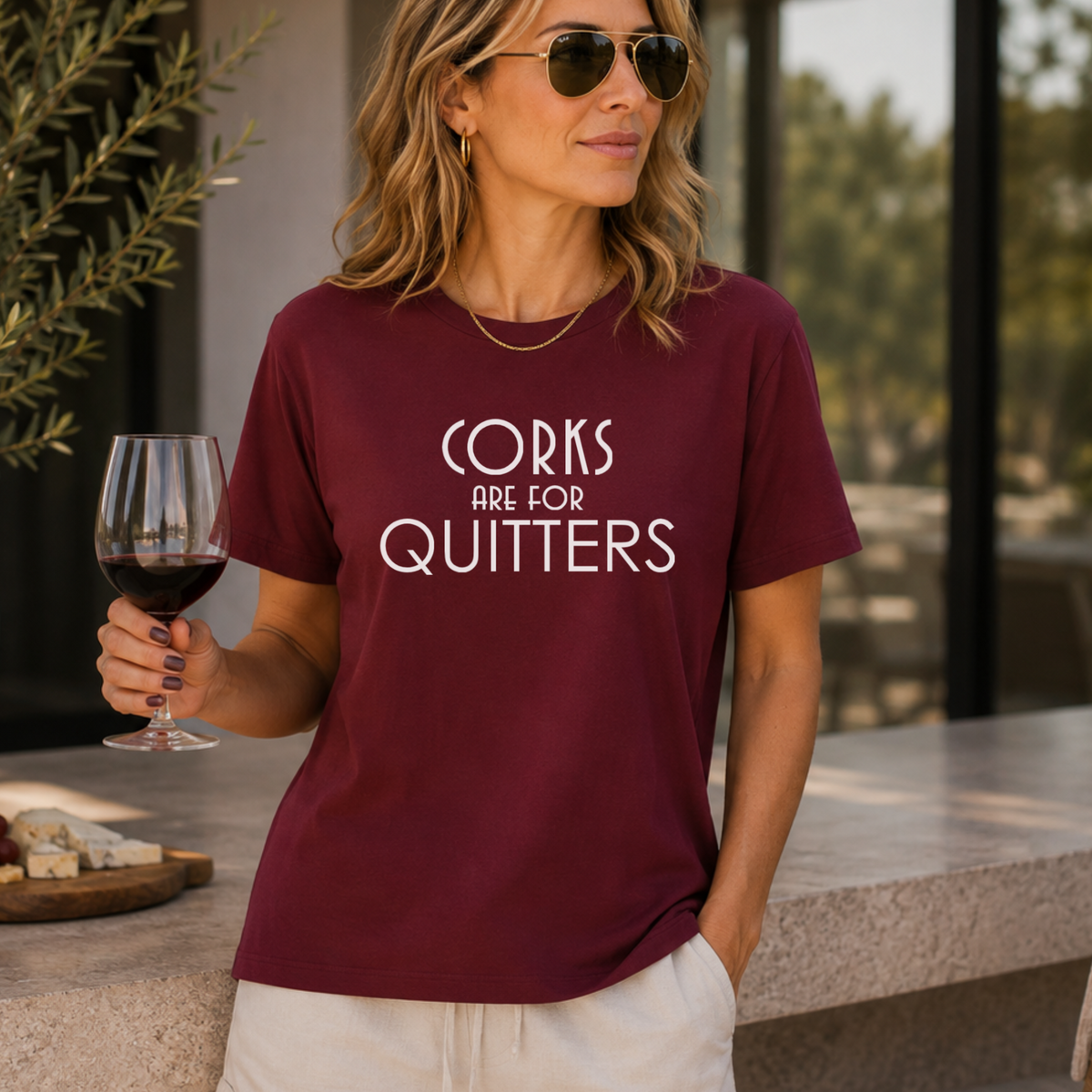A woman wearing a burgundy graphic t-shirt with the text 'CORKS ARE FOR QUITTERS' in white, paired with off-white shorts, holding a glass of red wine in a patio setting.