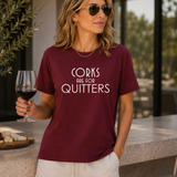 A woman wearing a burgundy graphic t-shirt with the text 'CORKS ARE FOR QUITTERS' in white, paired with off-white shorts, holding a glass of red wine in a patio setting.