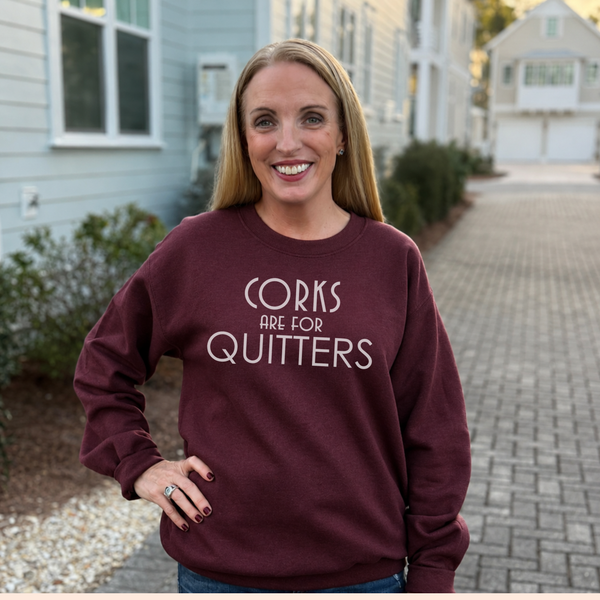A smiling woman stands outdoors on a paved driveway wearing a burgundy crewneck sweatshirt with the white text 'CORKS ARE FOR QUITTERS'.