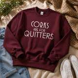A maroon crewneck sweatshirt with the text 'CORKS ARE FOR QUITTERS' in white, styled in a flat lay with jeans, white sneakers, a woven blanket, and green leaves.
