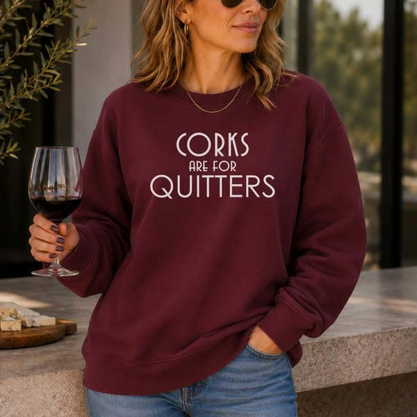 A woman wearing a maroon crewneck sweatshirt with the white text 'CORKS ARE FOR QUITTERS' stands in a patio setting holding a glass of red wine.