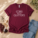 A maroon short-sleeve graphic T-shirt laid flat on a beige surface with wine-themed props, including a glass of red wine, grapes, corks, a corkscrew, and folded blue jeans. The shirt has white text reading “CORKS ARE FOR QUITTERS.