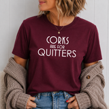 A woman wearing a maroon crewneck t-shirt with the white text 'CORKS ARE FOR QUITTERS' printed on the front, styled with a beige knit cardigan and blue jeans.