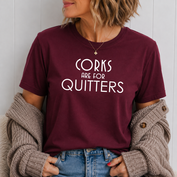 A woman wearing a maroon crewneck t-shirt with the white text 'CORKS ARE FOR QUITTERS' printed on the front, styled with a beige knit cardigan and blue jeans.