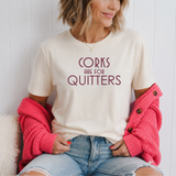 A woman wearing a cream-colored graphic t-shirt with the text 'CORKS ARE FOR QUITTERS' in dark red lettering, styled with a bright pink waffle-knit cardigan and light-wash denim shorts.