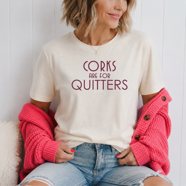 A woman wearing a cream-colored graphic t-shirt with the text 'CORKS ARE FOR QUITTERS' in dark red lettering, styled with a bright pink waffle-knit cardigan and light-wash denim shorts.