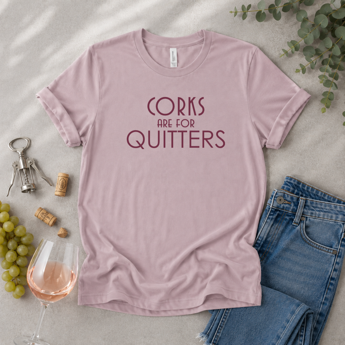 A dusty pink short-sleeve t-shirt with the text 'CORKS ARE FOR QUITTERS' printed in dark red across the chest, styled in a flat lay with jeans, a glass of rosé wine, grapes, a corkscrew, and eucalyptus leaves on a light gray textured surface.
