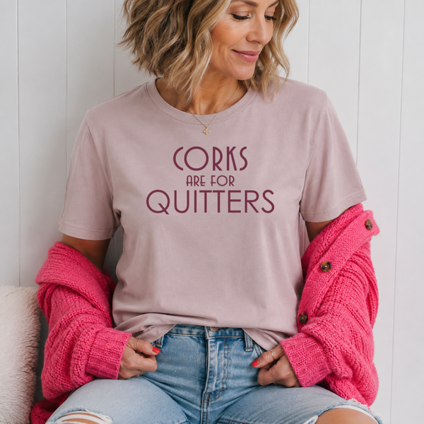 A woman wearing a dusty pink graphic t-shirt with the text 'CORKS ARE FOR QUITTERS' in dark maroon. She has a chunky bright pink knit cardigan draped off her shoulders and is wearing light-wash blue jeans, sitting against a white paneled wall.
