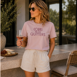 A woman wearing a light pink graphic t-shirt that reads 'CORKS ARE FOR QUITTERS' in dark purple text, paired with cream drawstring shorts, holding a glass of rosé wine in an outdoor patio setting.