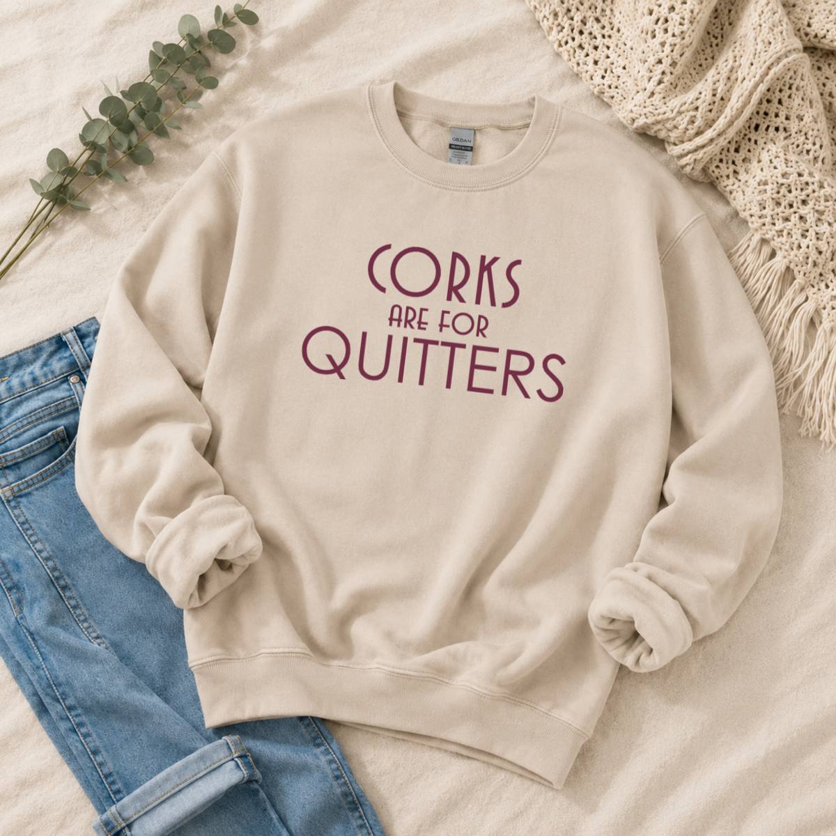 A beige crewneck sweatshirt with the text 'CORKS ARE FOR QUITTERS' printed in dark red on the front, styled in a flat lay with blue jeans, a green eucalyptus sprig, and a cream knit blanket.