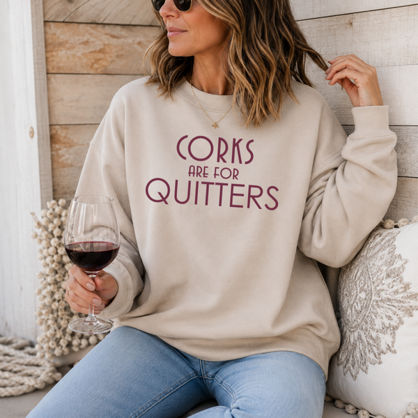 A woman wearing a beige crewneck sweatshirt with the text 'CORKS ARE FOR QUITTERS' printed in maroon across the chest, sitting in a rustic setting and holding a glass of red wine.