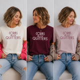 Three women seated side by side wearing the same slogan T-shirt in three colorways: cream, burgundy, and dusty pink. The shirts read 'CORKS ARE FOR QUITTERS' in dark lettering.