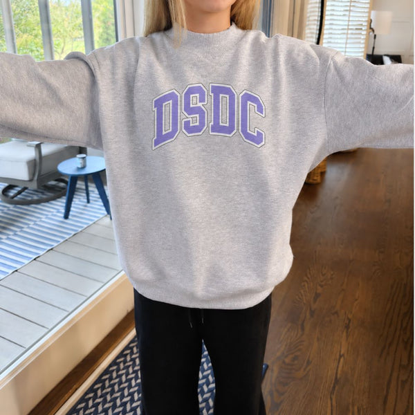 A person wearing a light heather gray crewneck sweatshirt with 'DSDC' printed across the chest in purple varsity-style lettering, standing indoors with arms outstretched.