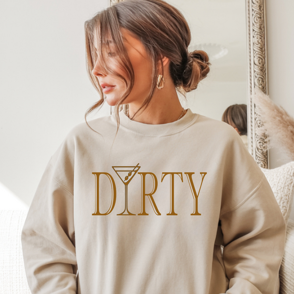 Woman wearing a beige sweatshirt with 'DIRTY' printed on it, sitting in a cozy room. The letter "i" in 'martini' is a martini glass.