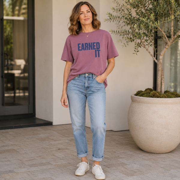 A woman standing outside in jeans and a berry colored Comfort Colors t-shirt that says "Earned It" in navy blue.